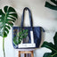 A blue tote bag painted with a mostera, and matching monstera background